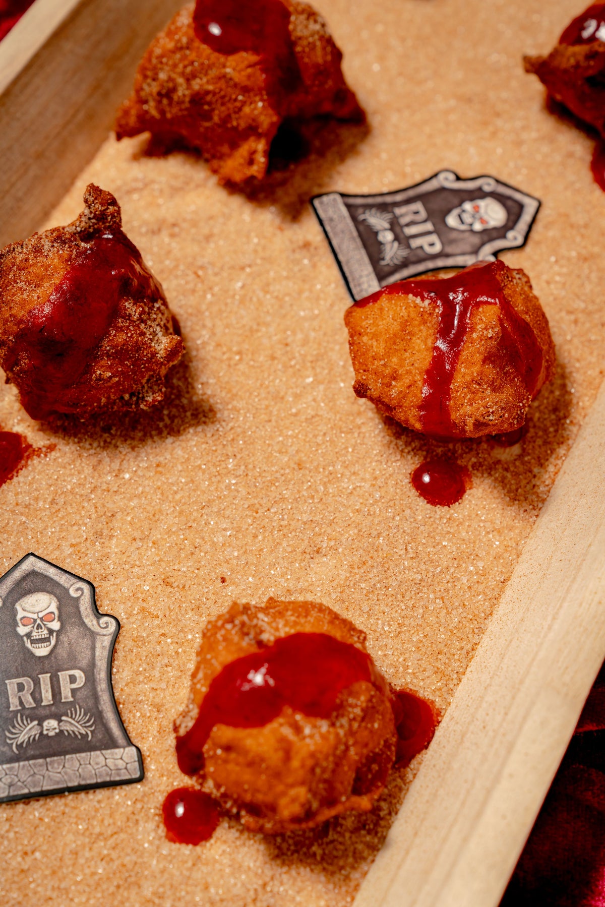 Bloody Good Apple Cider Donut Holes dusted with sugar, drizzled with cranberry blood glaze, and arranged on brown sugar with tombstone-shaped decorations.