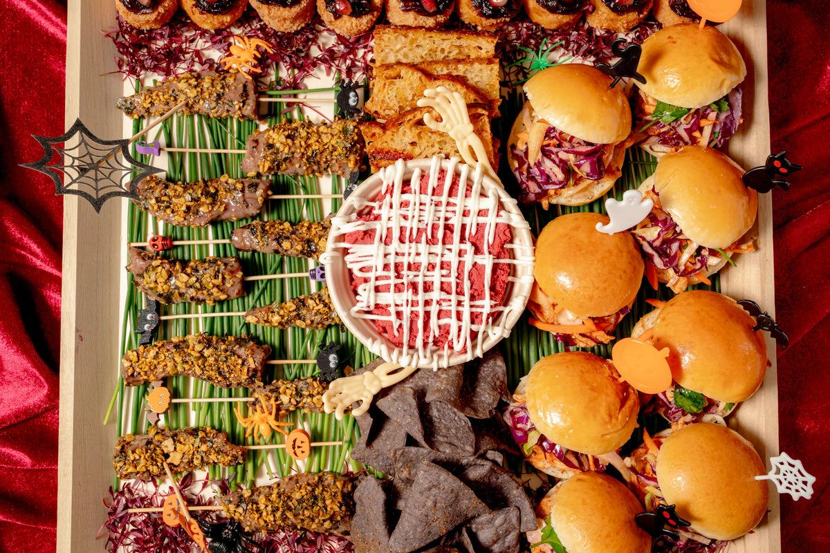 A Halloween Savory Platter featuring beef skewers, arancini, mini sandwiches, and black tortilla chips, with a central bowl of red dip drizzled in white and decorated with Halloween-themed toppers.