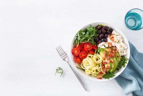 A bowl with spiralized zucchini, cherry tomatoes, olives, feta cheese, avocado, arugula, and bacon bits sits on a white table next to a fork, a blue napkin, and a glass of water.