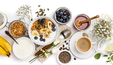 A breakfast spread with granola, banana, yogurt, blueberries, honey, chia seeds, coffee, and flowers on a white background.