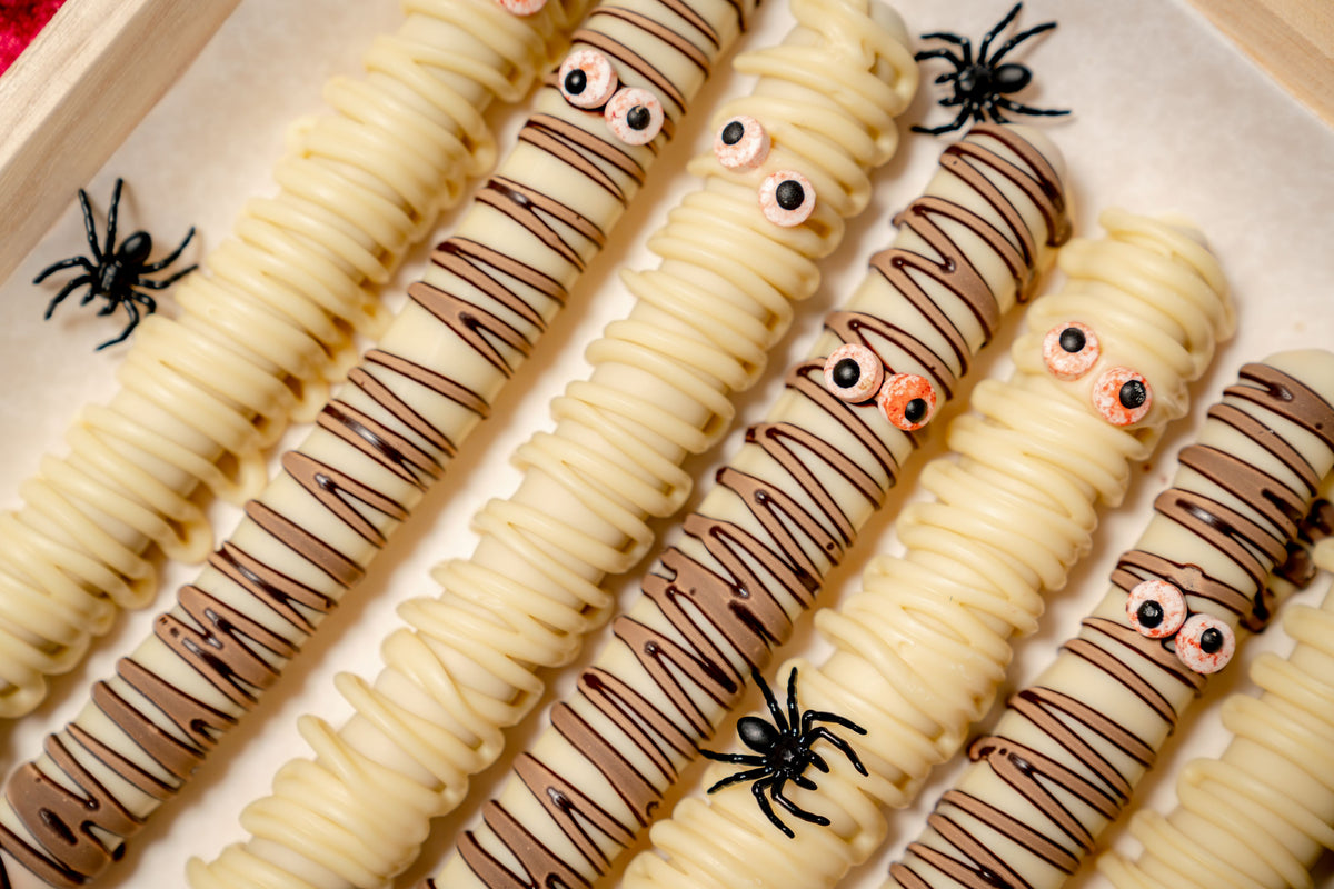 Pretzel Rod Ghosts, made of pretzel rods dipped in white chocolate and drizzled with milk chocolate to resemble mummies with candy eyes, decorated with small plastic spiders.