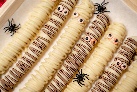 Pretzel Rod Ghosts, made of pretzel rods dipped in white chocolate and drizzled with milk chocolate to resemble mummies with candy eyes, decorated with small plastic spiders.