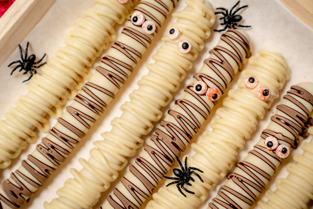 Pretzel Rod Ghosts, made of pretzel rods dipped in white chocolate and drizzled with milk chocolate to resemble mummies with candy eyes, decorated with small plastic spiders.