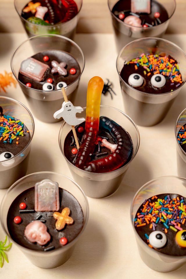 Tombstone Shooters consisting of small plastic cups filled with chocolate pudding, cookies & cream crumble, gummy worms, candy tombstones, googly eyes, and colorful Halloween sprinkles.