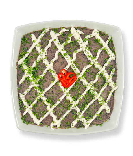 A square dish of 6 Layer Dip topped with Whitestone Cheddar, a mayonnaise drizzle, green onions, and a red chili pepper slice in the center. Serve with guacamole and corn tortillas for a complete flavor experience.