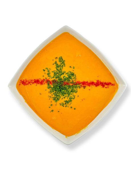 In a square white bowl, the NY Beer Cheese Dip cradles orange soup adorned with a stripe of green herbs and red seasoning, echoing the vibrant hues of Whitestone Cheddar.