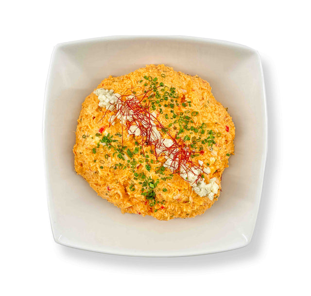 A vibrant orange Buffalo Chicken Dip in a square white bowl is garnished with crumbled cheese, chopped chives, and fine red threads, making it the perfect pairing for crispy corn tortillas.
