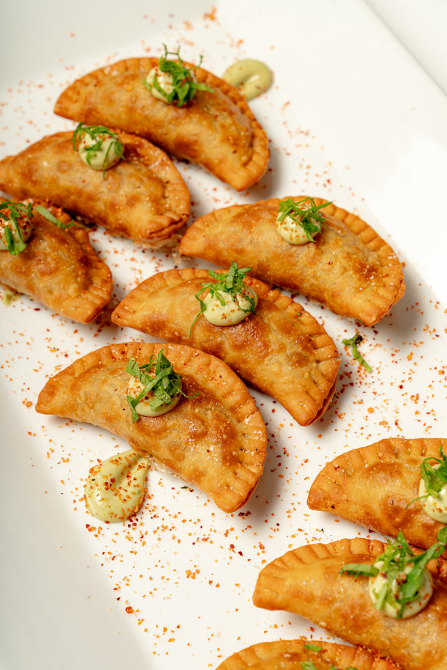 Eight Mini Vegetable Empanadas are arranged on a white plate, each topped with vegan aioli and finished with green herbs and a sprinkle of Tajin seasoning.