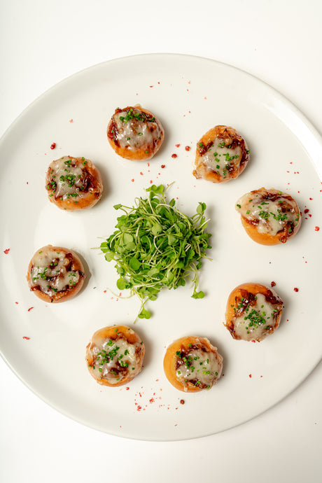 Eight French Onion Soup Boules, topped with Gruyere, caramelized onions, and herbs, are arranged in a circle on a white plate with microgreens in the center and a sprinkle of red seasoning around.