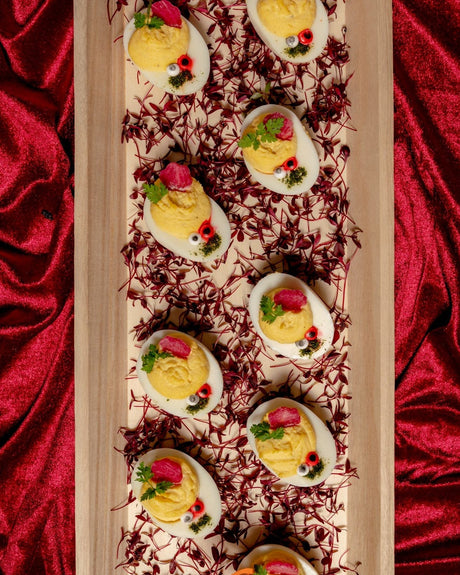 Devilish Eggs decorated with edible eyes, herbs, and red vegetable pieces on a bed of microgreens, served on a rectangular wooden platter.