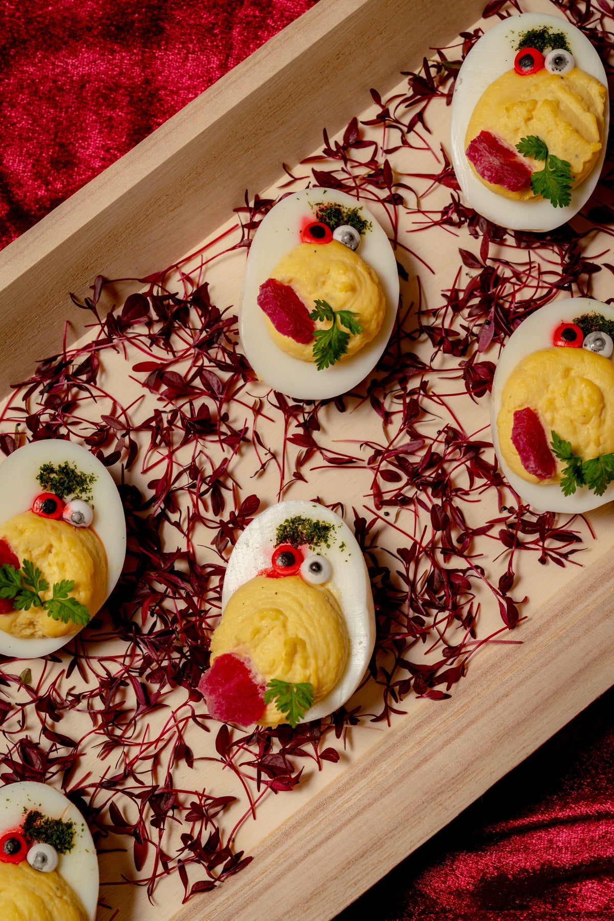 Devilish Eggs decorated as small creatures with candy eyes, beet slices, and herbs, arranged on a spooky spider web bed of red microgreens in a wooden tray.