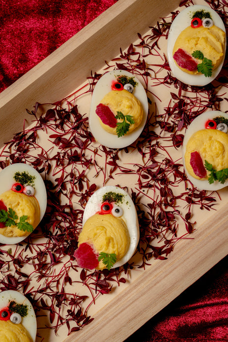 Devilish Eggs decorated as small creatures with candy eyes, beet slices, and herbs, arranged on a spooky spider web bed of red microgreens in a wooden tray.