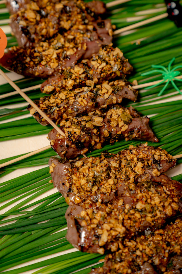 Witch’s Stake Beef Skewers glazed in pomegranate, topped with crushed nuts and herbs, arranged on a bed of green chives with small plastic spider decorations.
