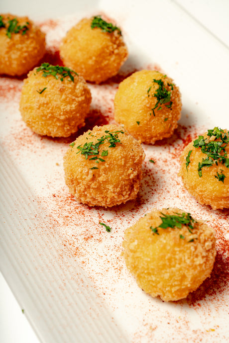 Six golden Mac N Cheese Croquettes with aged cheddar, coated in a herbed crust and sprinkled with red seasoning, arranged on a white plate with chopped herbs.