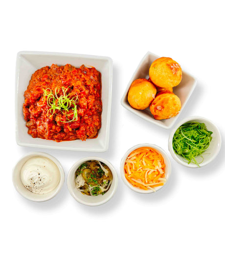A square dish features reddish stew of aged beef chili, topped with green garnish. Accompanying it are round bread rolls in another dish, along with small bowls of sour cream, sautéed veggies, shredded cheese, and fresh greens—all arranged elegantly on a white background from Mom's Chili Bar.