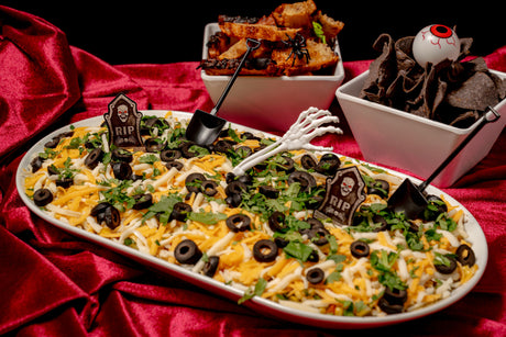 A 6 Feet Deep Graveyard Dip Platter featuring a rectangular 6-layer dip topped with black olive slices, cilantro, tombstone signs, and a plastic skeleton hand, served with toasted bread and chips.