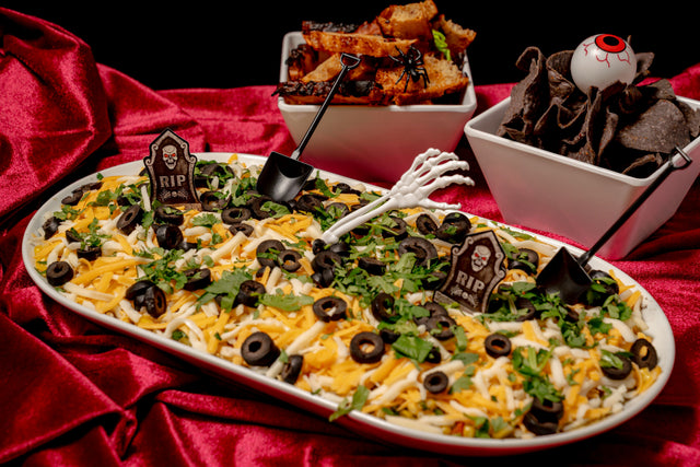 A 6 Feet Deep Graveyard Dip Platter featuring a rectangular 6-layer dip topped with black olive slices, cilantro, tombstone signs, and a plastic skeleton hand, served with toasted bread and chips.