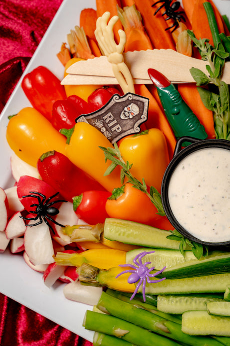 A Creepy Crudité Platter of rainbow veggies like bell peppers, cucumber, and radishes, decorated with plastic spiders, a skeleton hand, and a tombstone sign, served with a bowl of dip.