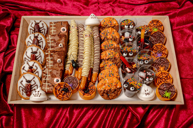 The Haunted Bake Shop Platter is a wooden tray filled with pumpkin spice cookies, frosted chocolate brownies, pretzel rods, and pudding cups topped with candy eyes and gummy worms.