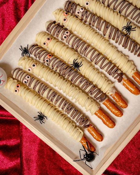 Pretzel Rod Ghosts made of white chocolate-dipped pretzel rods drizzled with icing to look like mummies with candy eyes, arranged in a wooden tray with small plastic spiders.