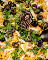 A 6 Feet Deep Graveyard Dip Platter featuring a 6-layer dip topped with shredded cheese, sliced black olives, chopped parsley, and a tombstone decoration with a skull and the letters “RIP.”