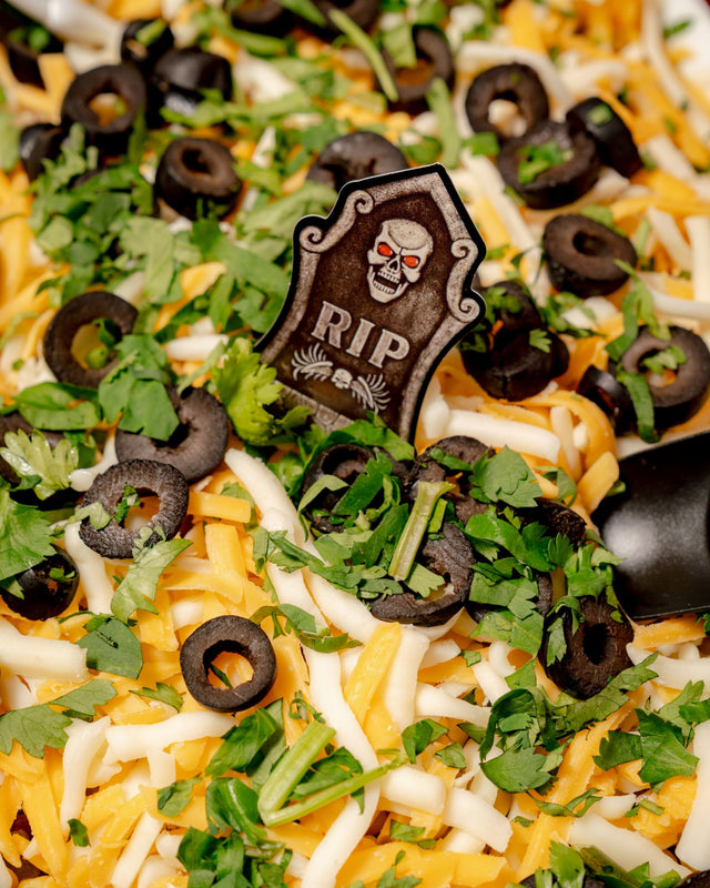 A 6 Feet Deep Graveyard Dip Platter featuring a 6-layer dip topped with shredded cheese, sliced black olives, chopped parsley, and a tombstone decoration with a skull and the letters “RIP.”
