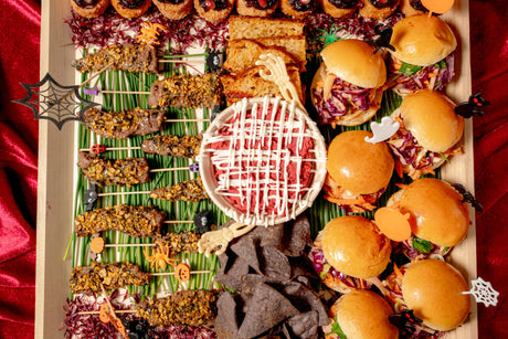 A Halloween Savory Platter featuring beef skewers, arancini, mini sandwiches, and black tortilla chips, with a central bowl of red dip drizzled in white and decorated with Halloween-themed toppers.