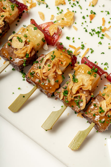 Four Short Rib Kebab skewers of grilled meat and vegetables, topped with crispy fried onions, chopped chives, and tangy pickled red onion, arranged on a white plate.