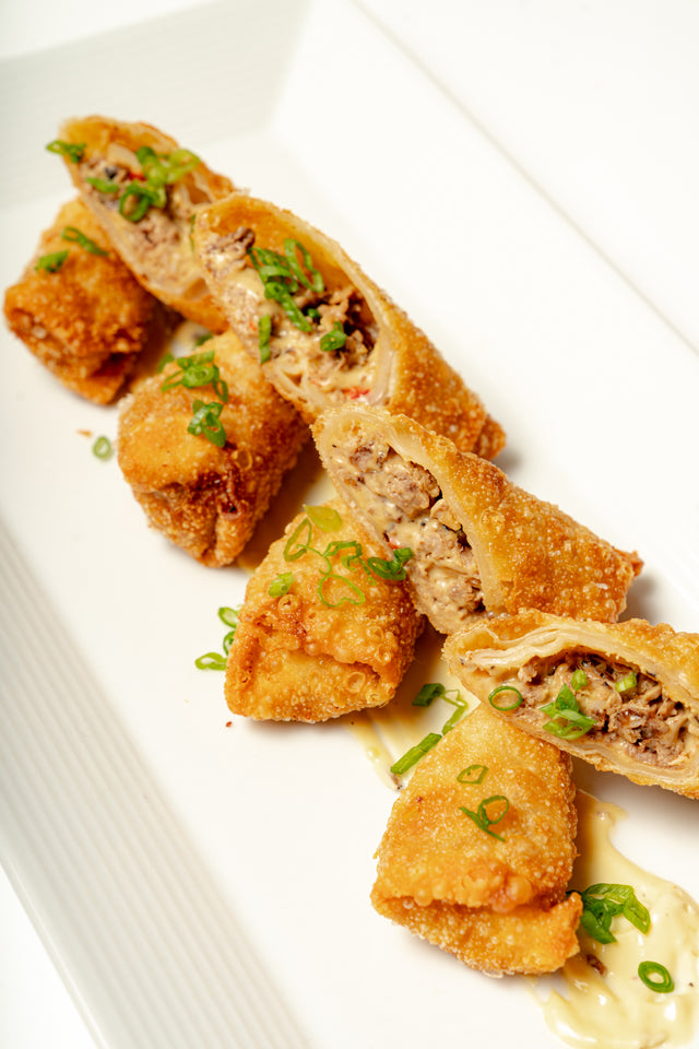 Truffled Cheesesteak Egg Roll: Crispy egg rolls filled with shaved ribeye, cut in half, topped with green onions, and served on a white plate with a drizzle of fondue dipping sauce.