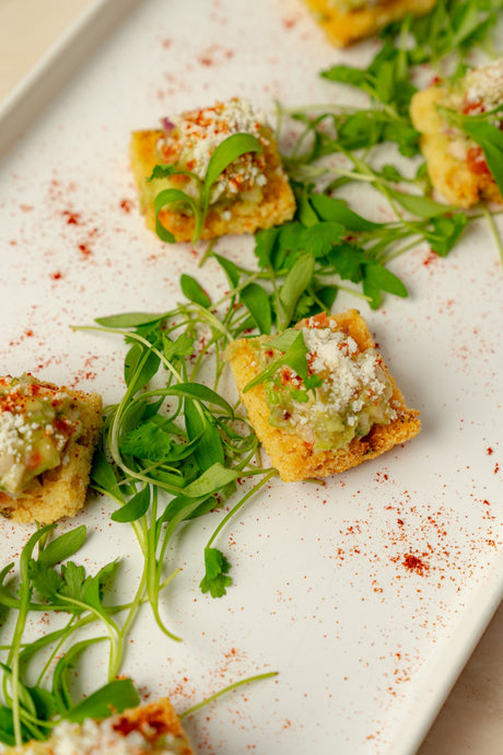 Deconstructed Chicken Guacamole is served on a white plate with fresh greens, topped with avocado, Cotija cheese, herbs, and a light dusting of red spice.