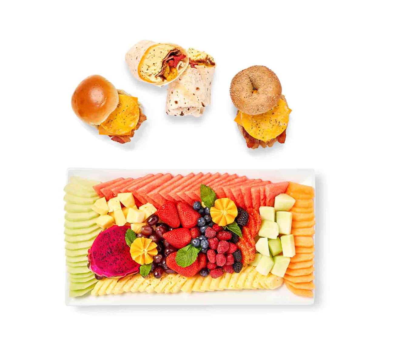A colorful fruit platter with melon, pineapple, and berries sits invitingly. Above it lie two Mini Breakfast Sandwiches and a bacon-cheese Wrap. Ideal when paired with your favorite morning beverages for a delightful start to the day.