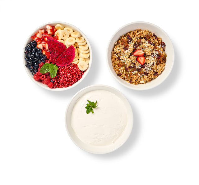 The Make Your Own Yogurt, Granola and Fruit Bar features three white bowls—one with seasonal fruit, one with homemade granola and strawberries, and one with plain yogurt topped with mint—arranged in a triangle on a white background.
