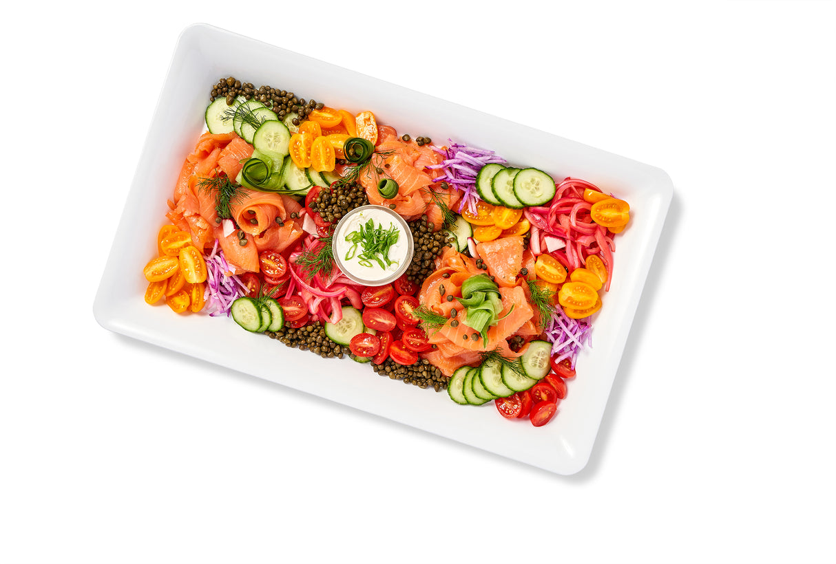 A white plate with a variety of vegetables, Nova Lox & Bagels Platter and capers on it.