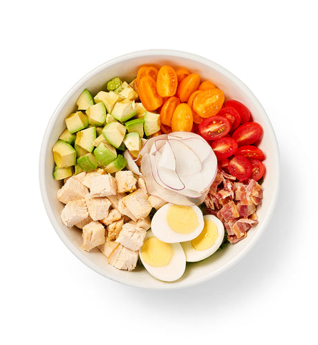 A bowl filled with Cobb Salad, avocado, tomatoes and eggs topped with crispy bacon.